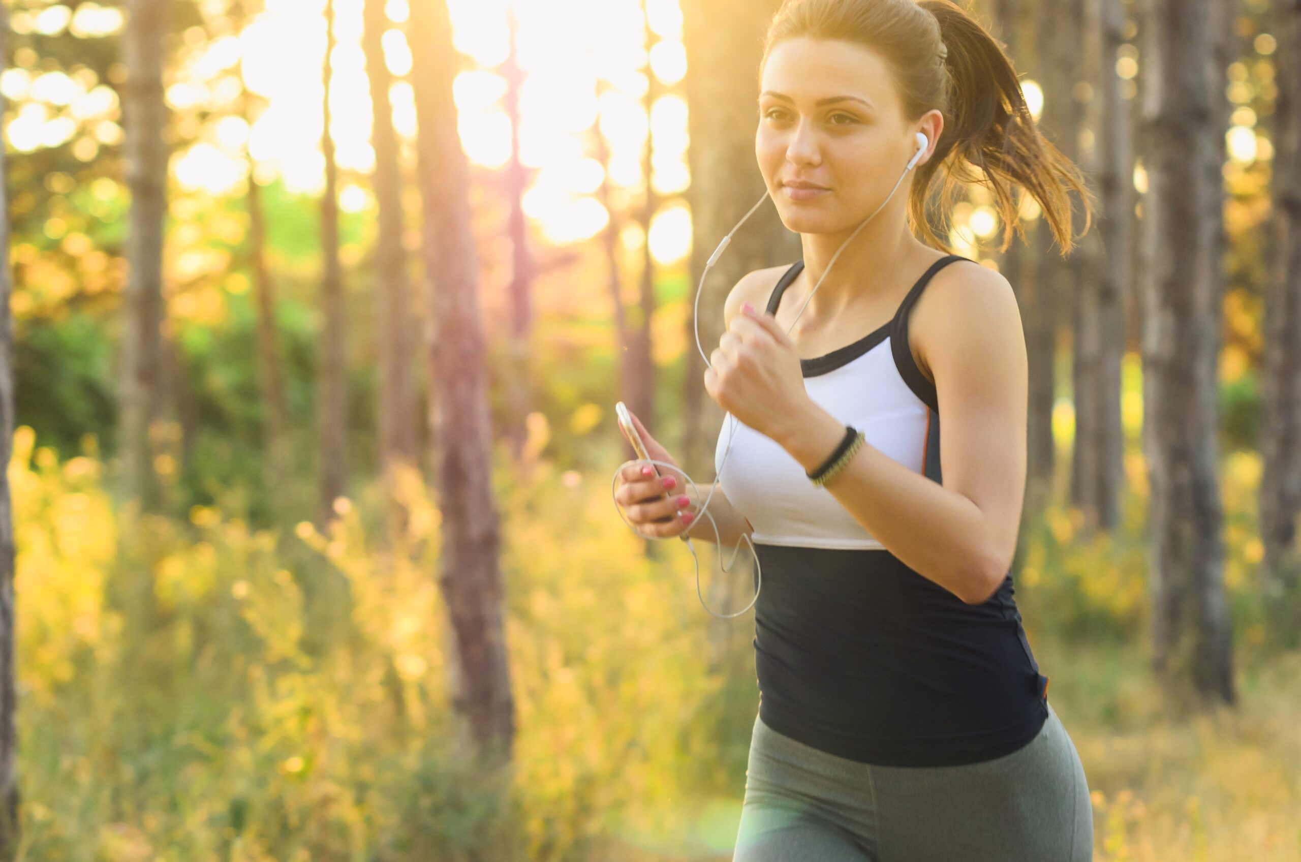 Woman jogging through a sunlit forest trail with earphones — running outdoors burns around 600 calories per hour, reduces cortisol levels and boosts mood through natural endorphin release, making it one of the best fat burning exercises for women